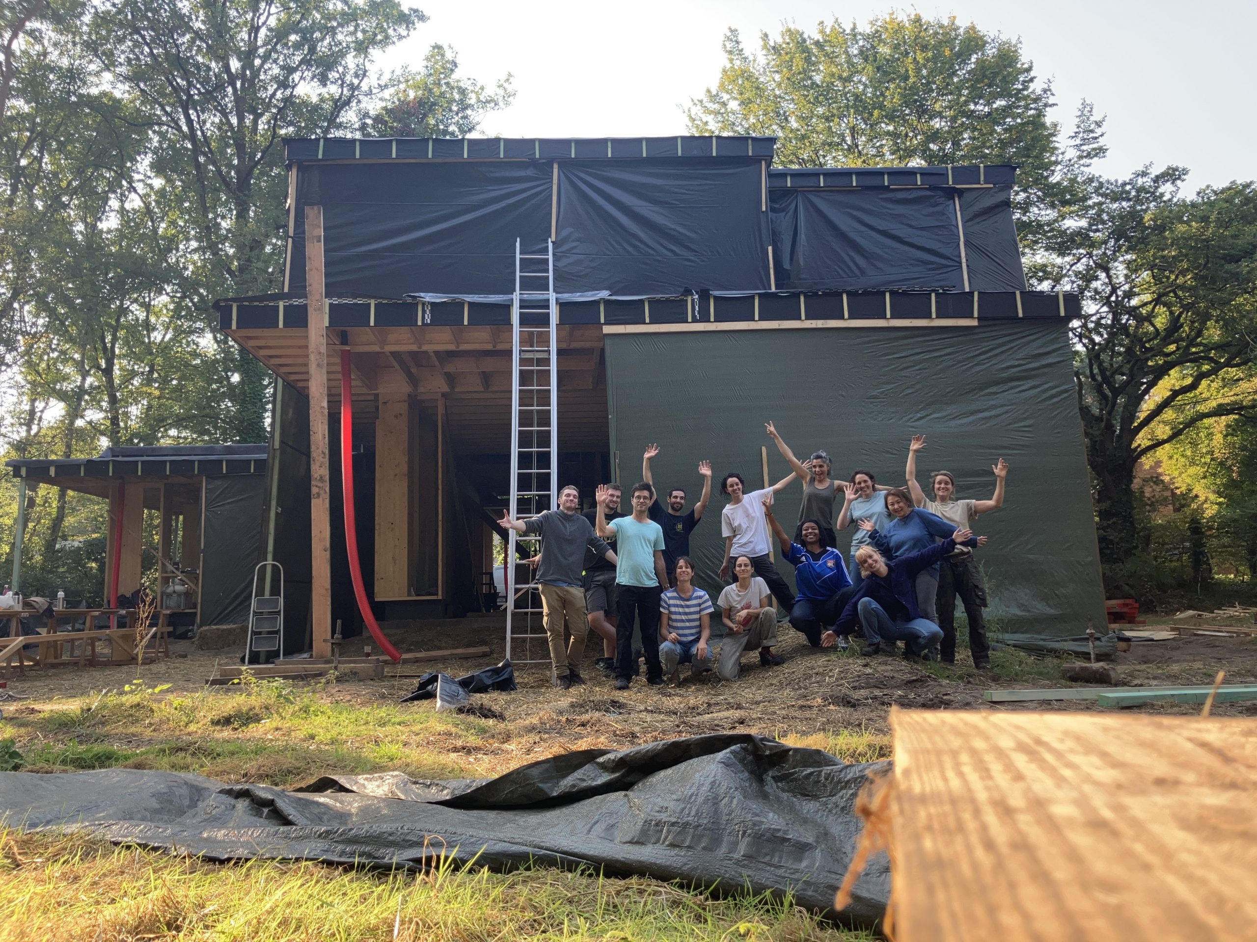 Une quinzaine de bénévoles, bras levés, posent devant une maison en ossature bois enveloppée d'un pare-pluie noir, lors d'un chantier participatif.