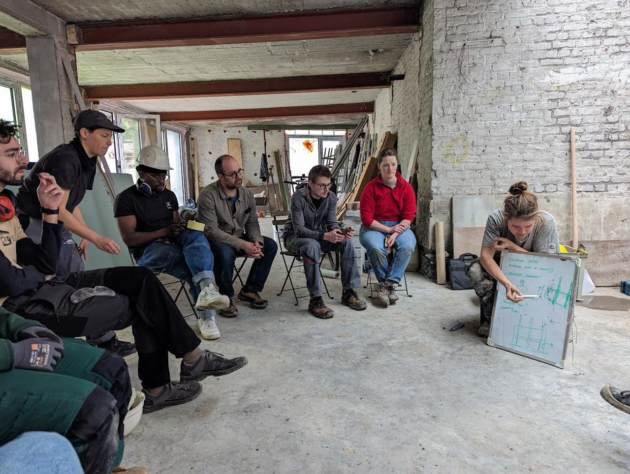 Laurane, accroupie devant un tableau blanc couvert d'un schéma technique, explique une étape à un groupe de participants assis sur un chantier aux murs de brique.