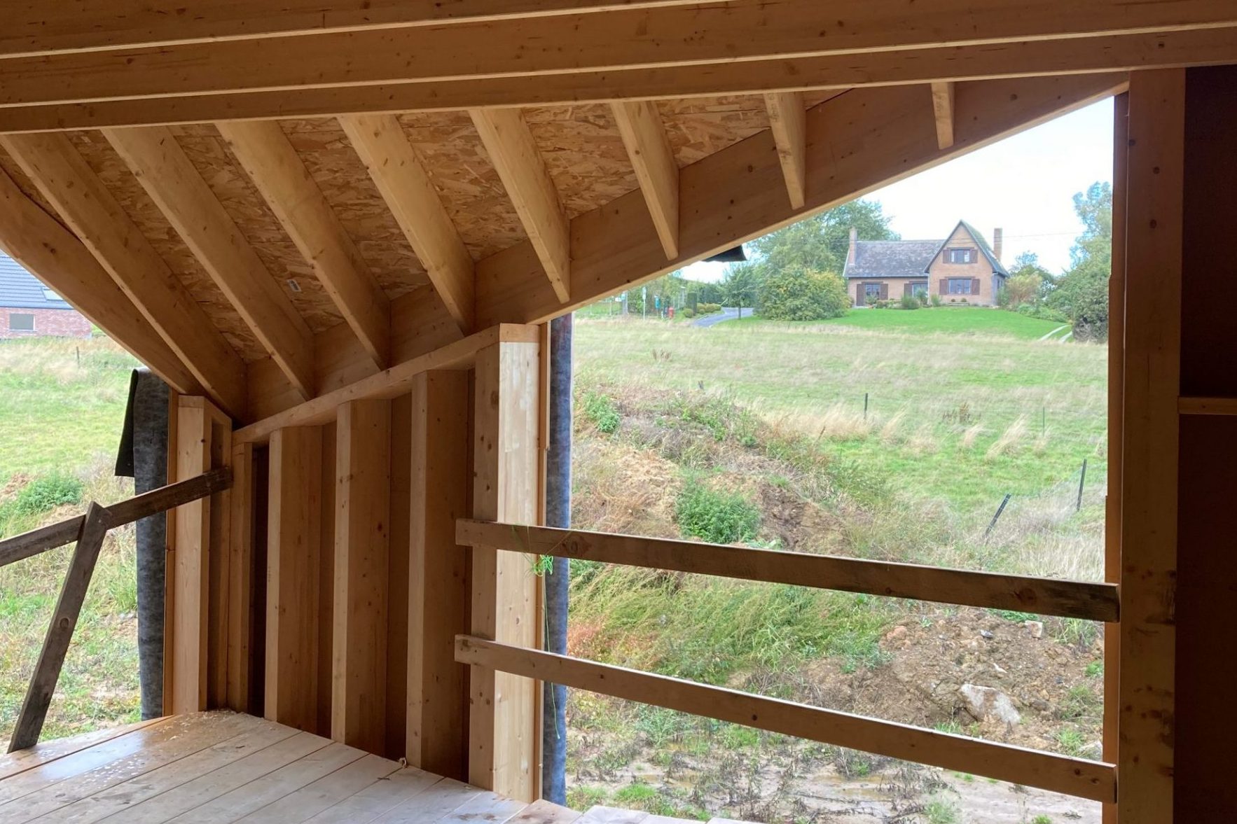 Vue depuis l'intérieur d'une ossature bois vers la campagne : charpente et montants clairs encadrent un pré vert et une maison sur la colline.