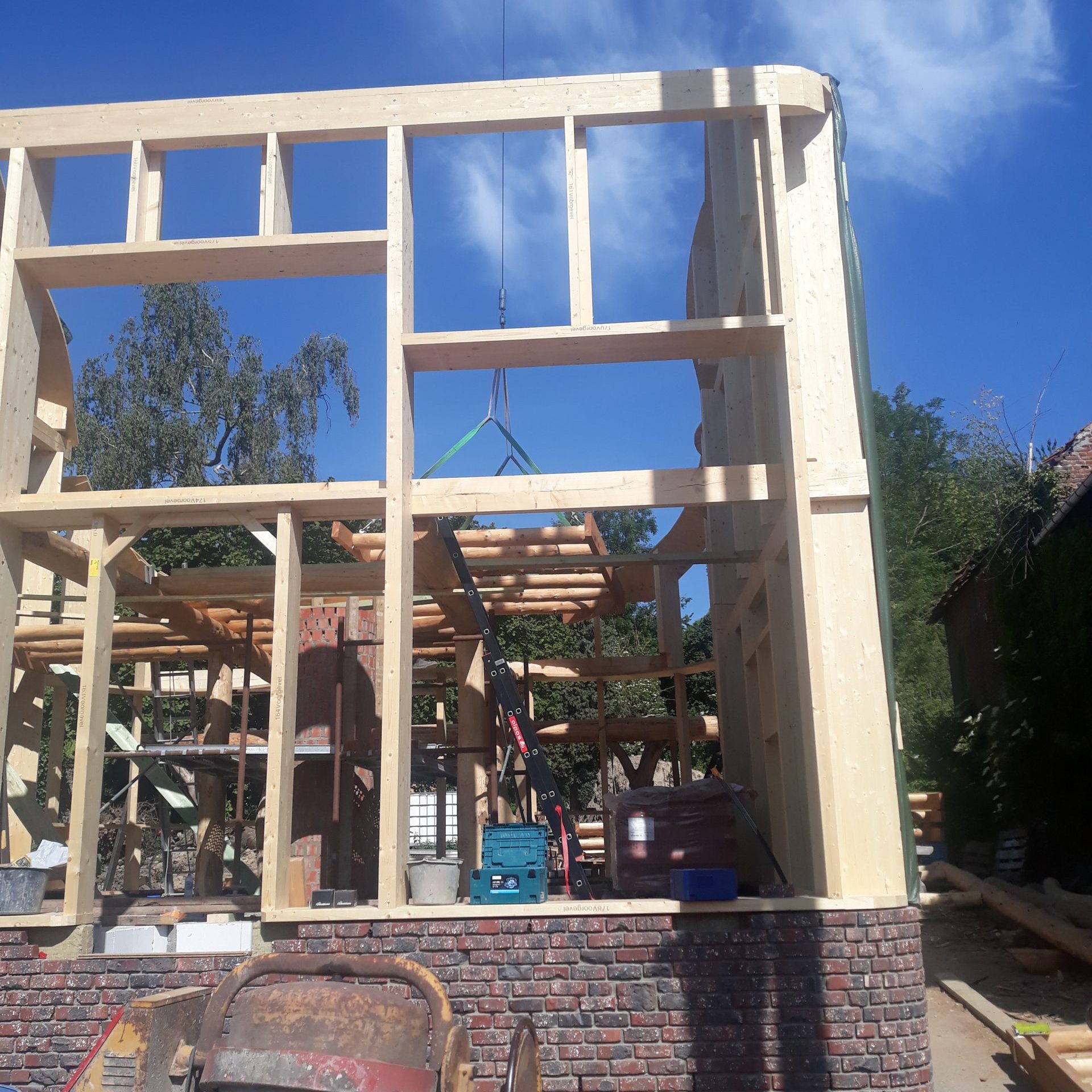 Ossature en bois d'une maison en cours de construction, montants et poutres clairs se détachant sur un ciel bleu, au-dessus d'un soubassement de briques.