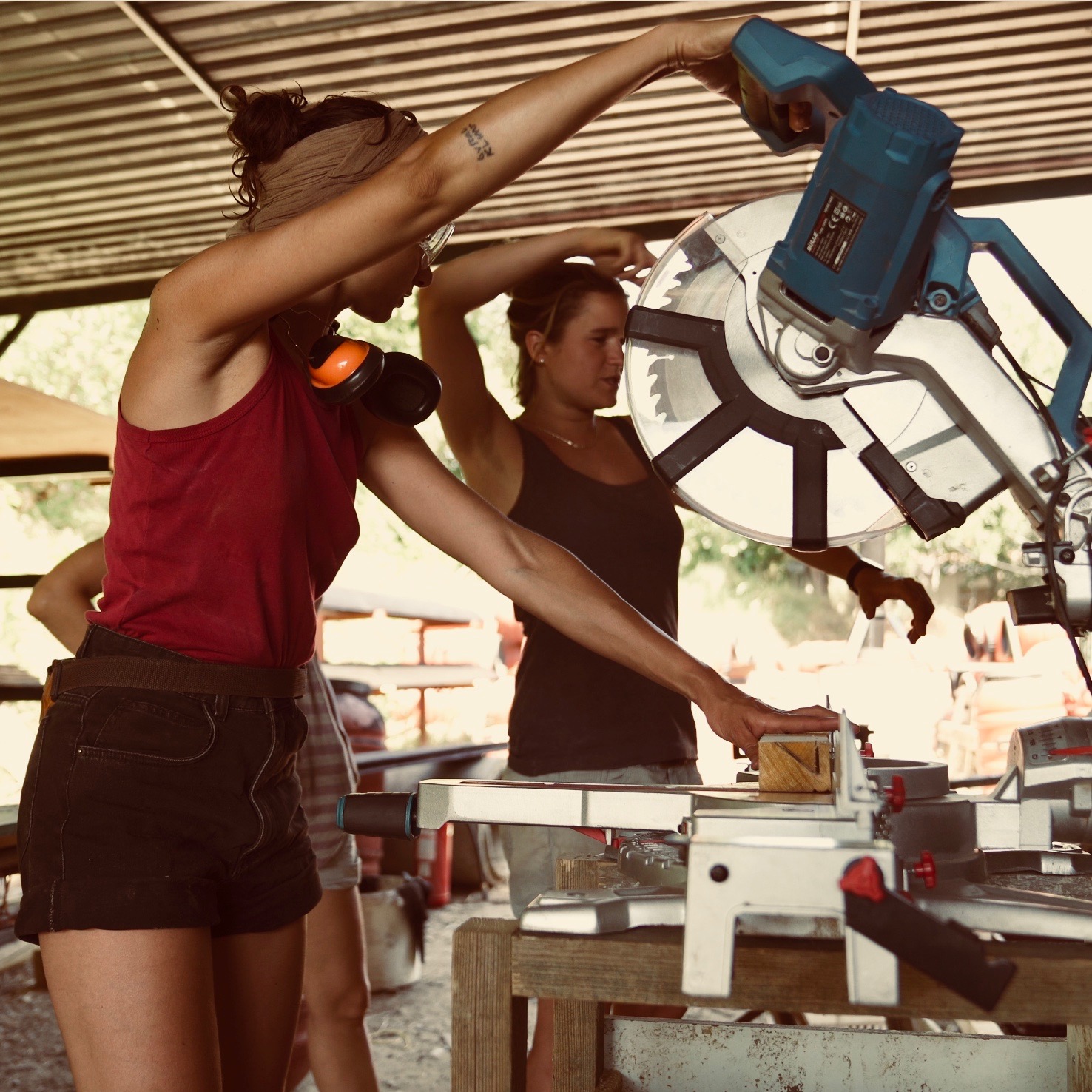 Deux femmes travaillent ensemble à une scie à onglet dans un atelier, l'une guidant la planche, l'autre observant la coupe.