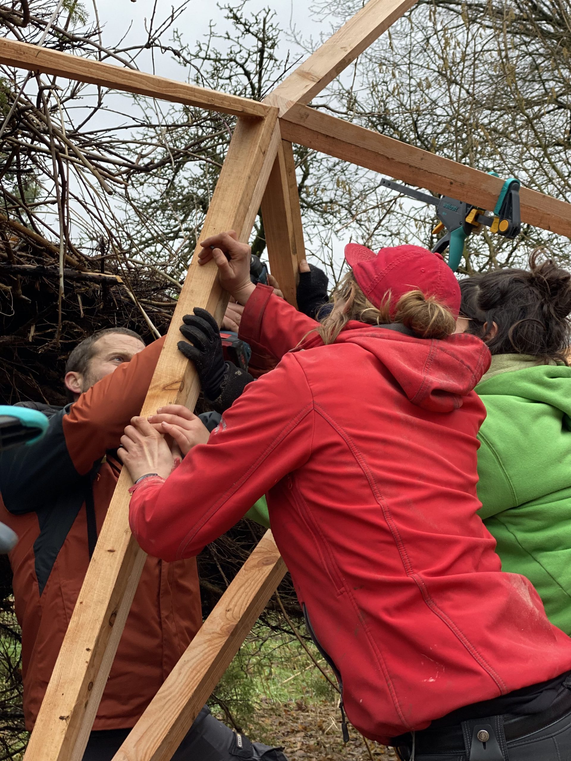 Un groupe soulève ensemble une structure de bois triangulée, chacun soutenant une partie du cadre à bout de bras.