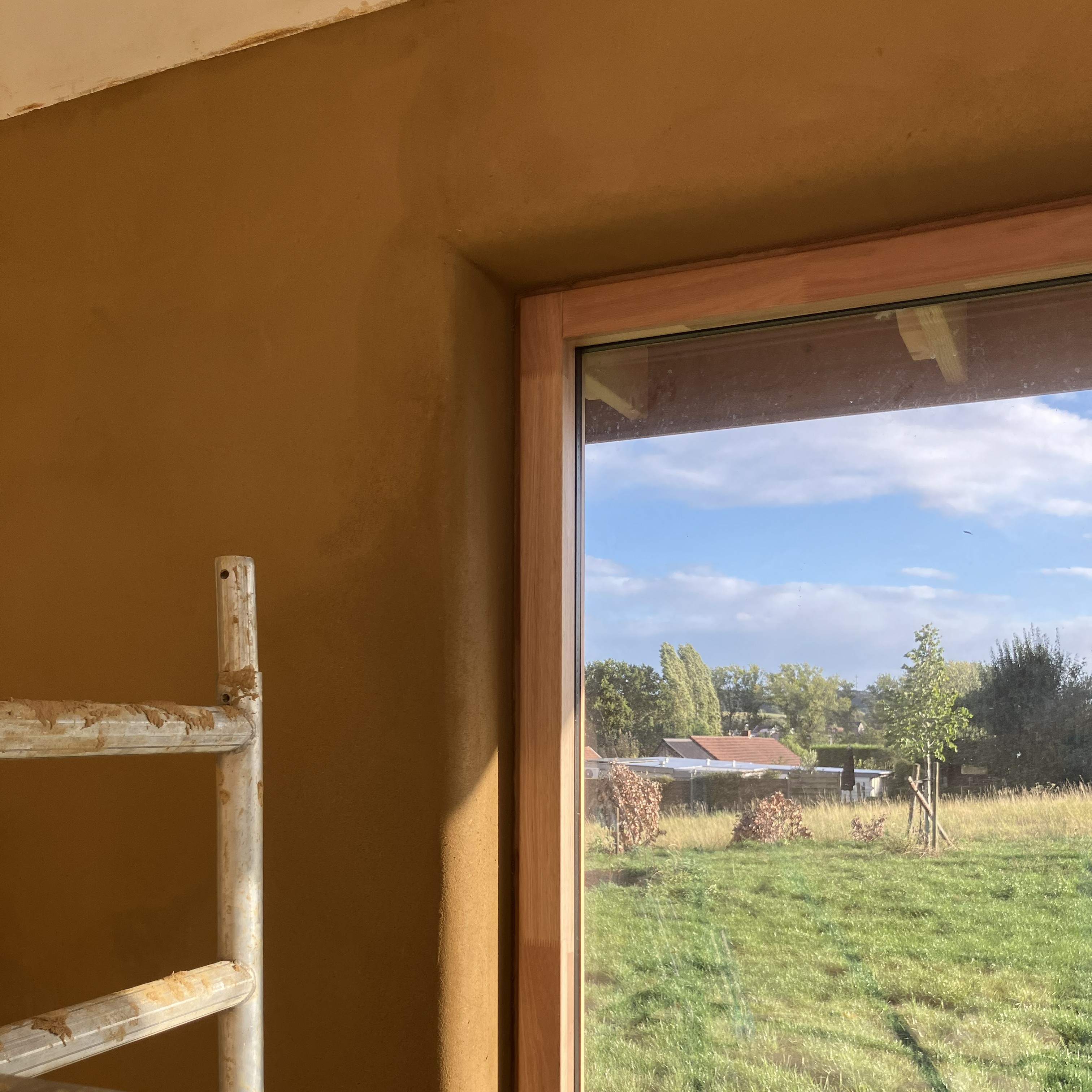 Angle d’une pièce en enduit terre ocre, linteau et fenêtre ouvrant sur la campagne