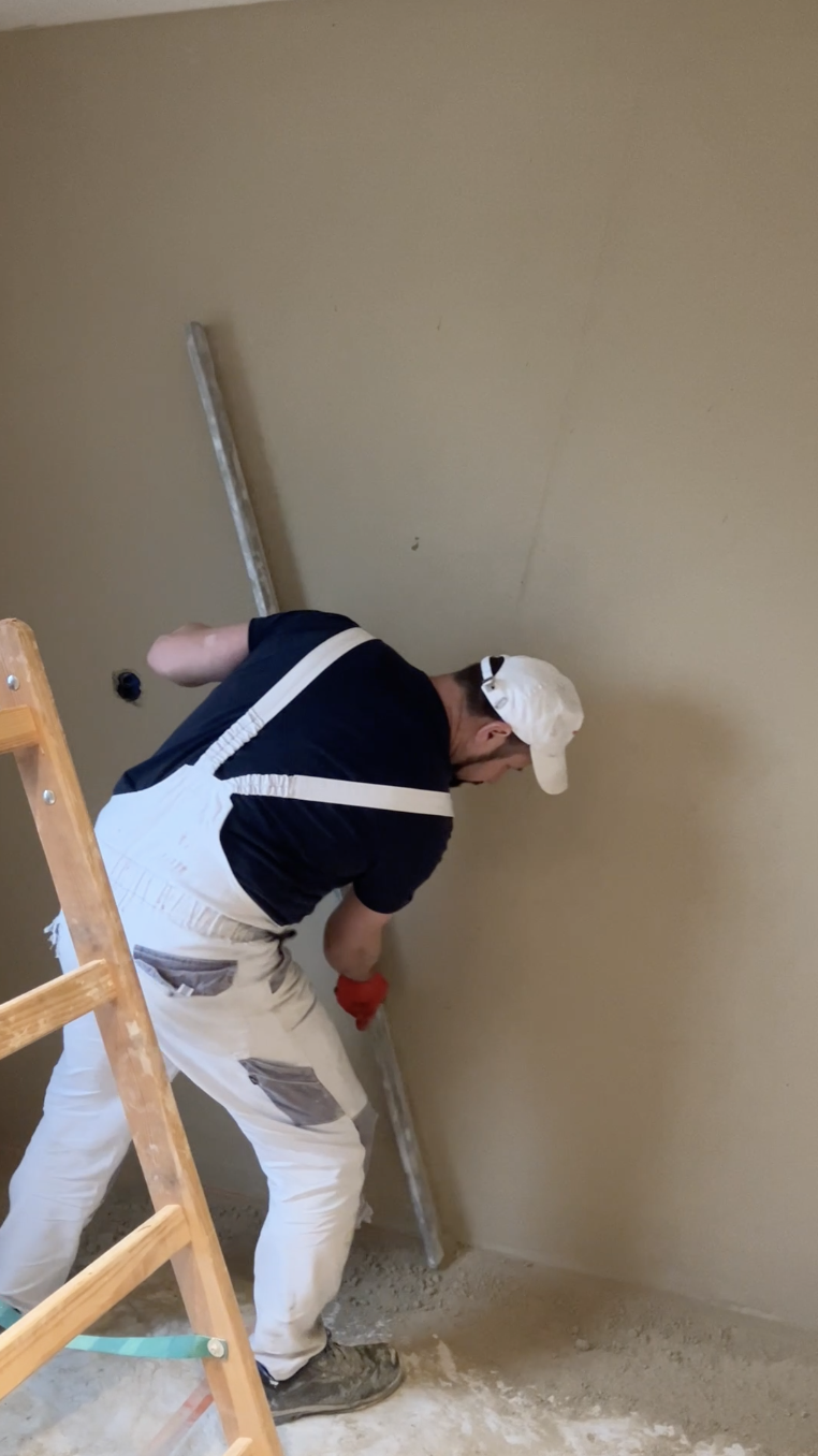 Un participant casqué lisse le haut d'un mur à la chaux, debout sur un escabeau