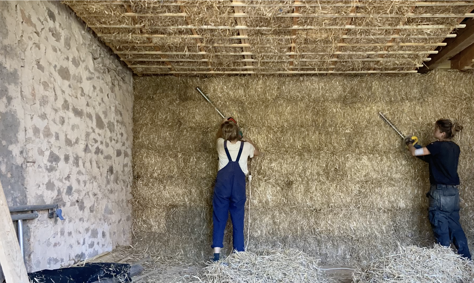 Deux participants enduisent un grand mur en paille sous la charpente